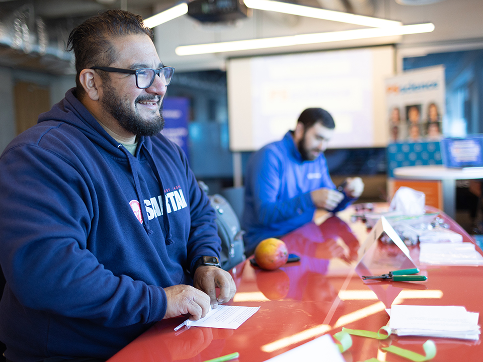 One of Telesign's team members smiling as he works on a project at a table. Another Telesign employee is working on a project in the background at the same table.