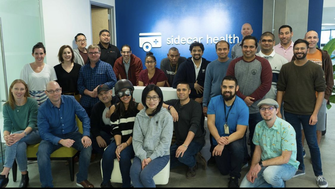 Sidecar Health group photo in the office with the compnay logo on a blue wall behind them