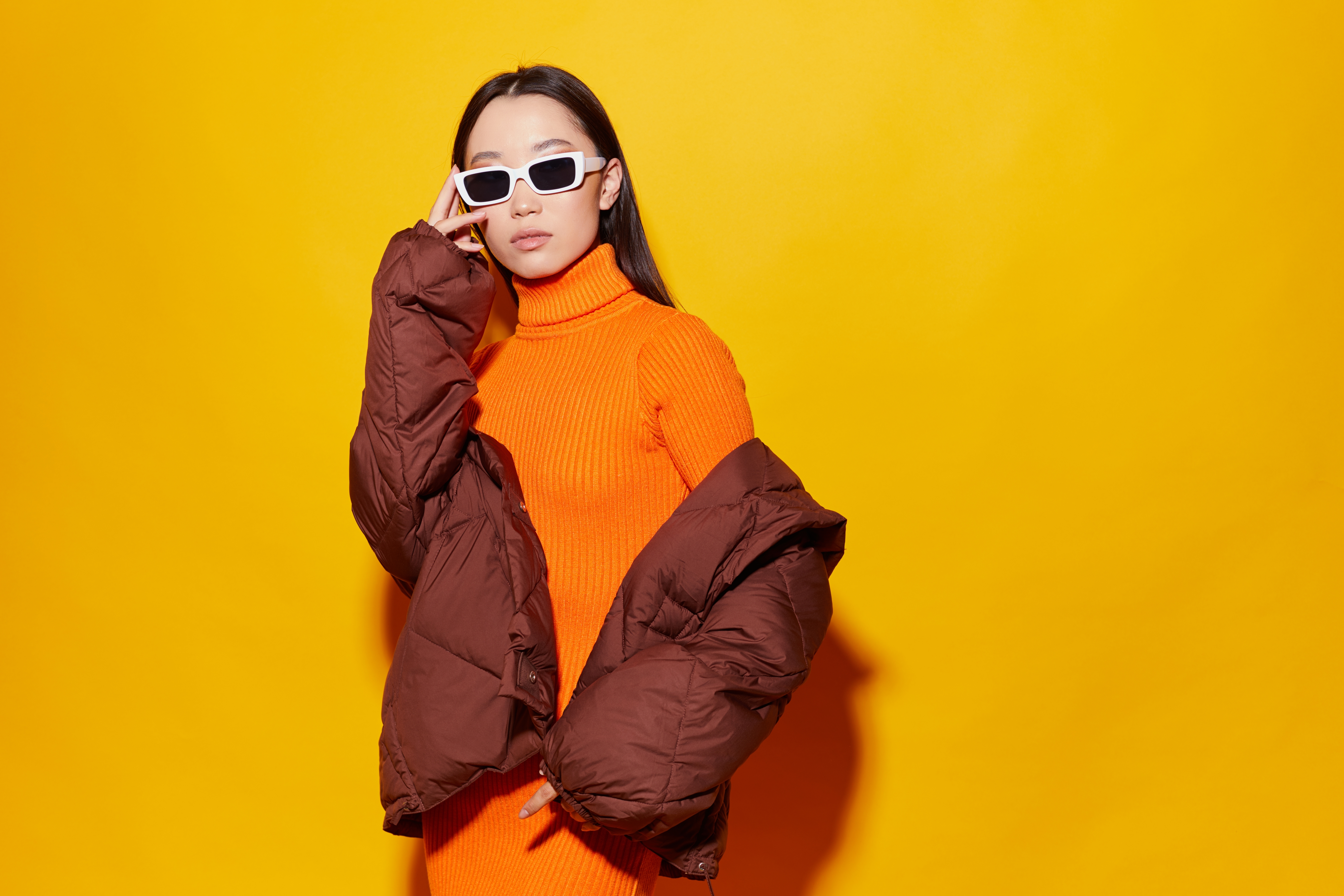 Stock photo of a young Asian woman in stylish clothes and sunglasses in front of a sunny yellow background.