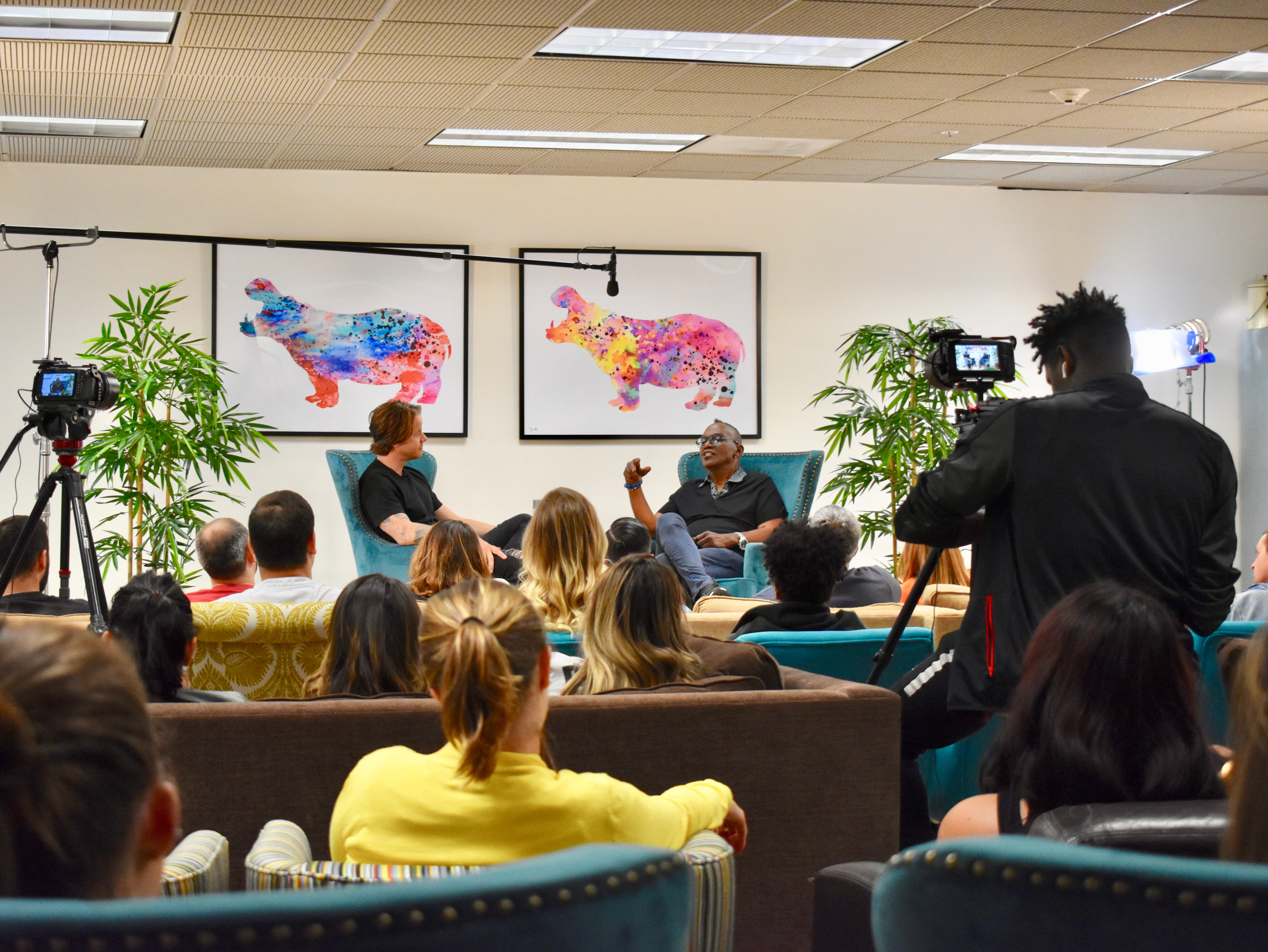 A fireside chat event with two leaders in armchairs talking in front of an audience of team members and cameras. 