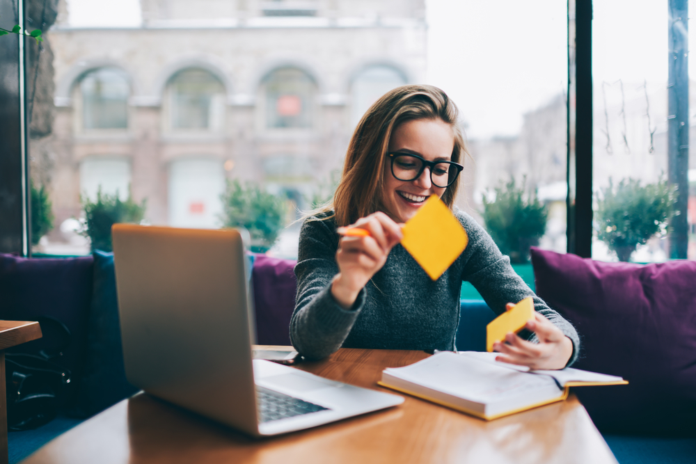 Woman working from coffee shop near window, laptop open, smiling as she tears off sticky notes from her to-do list