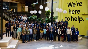  Large group photo of GoodRx team members in their office, standing near a staircase and a two-story yellow wall with the words “We’re here to help.”