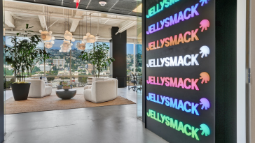 jellysmack office the logo runs up and down the wall to the right and we can see a beautifully designed seating area in the background with white furniture and indoor plants