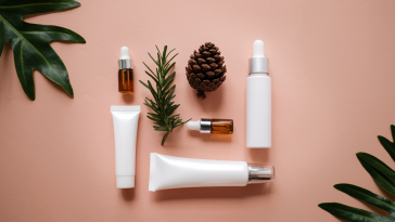 Peach background with fern leaves in the corners. Various tubes of beauty products are organized in a square in the middle.